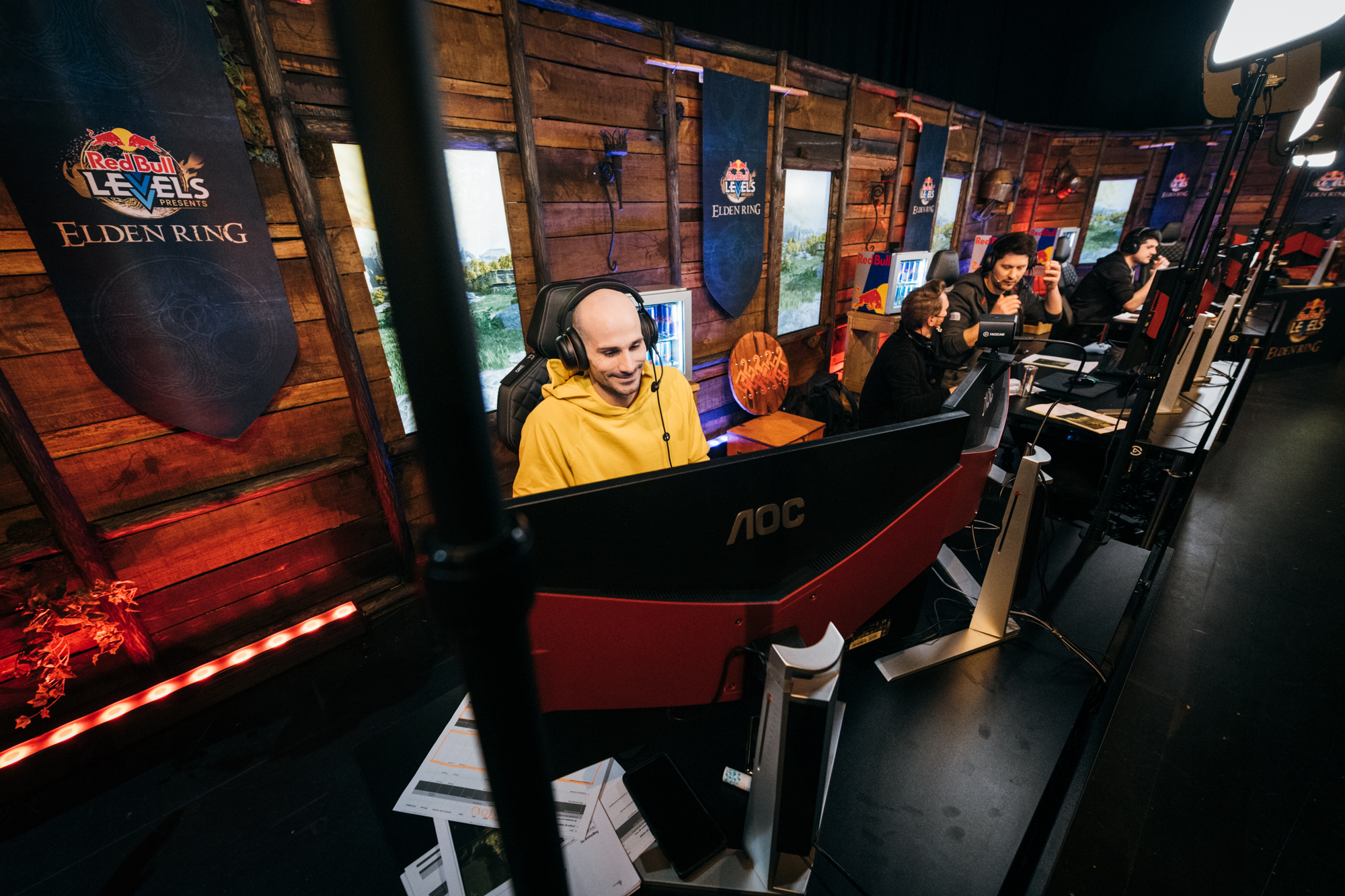 A group of gamers sits at computer desks in a wooden, decorated room with “Red Bull Levels Elden Ring” banners, focused on their screens. One player in front, part of the Elden Ring global XR event, wears a yellow hoodie and headset.