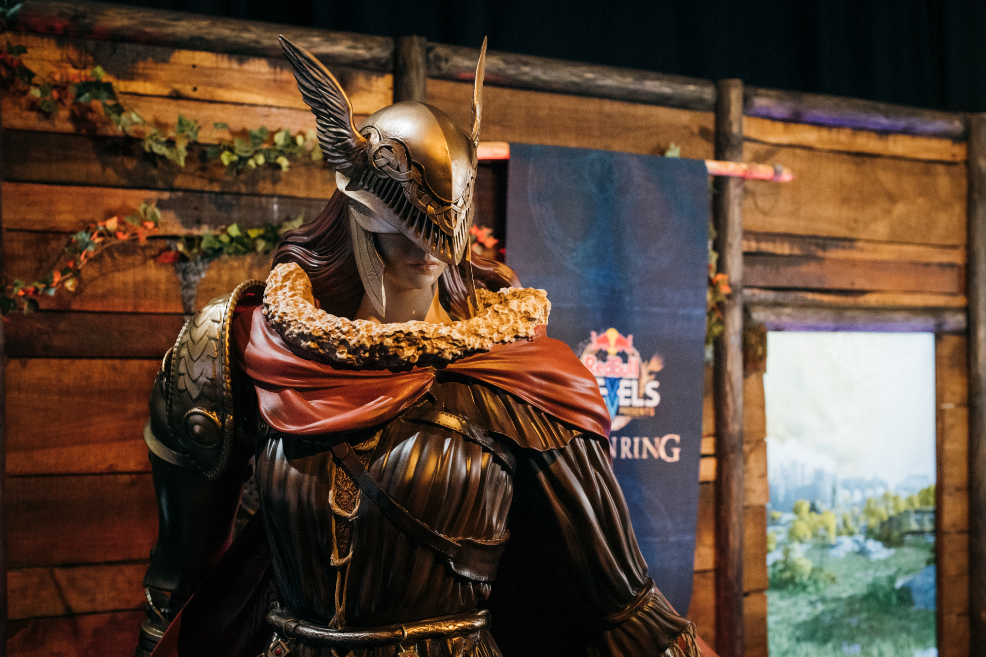 A detailed statue of a fantasy armored warrior, reminiscent of Elden Ring, with a winged helmet and fur-trimmed cloak stands in front of a wooden backdrop decorated with leaves and banners.