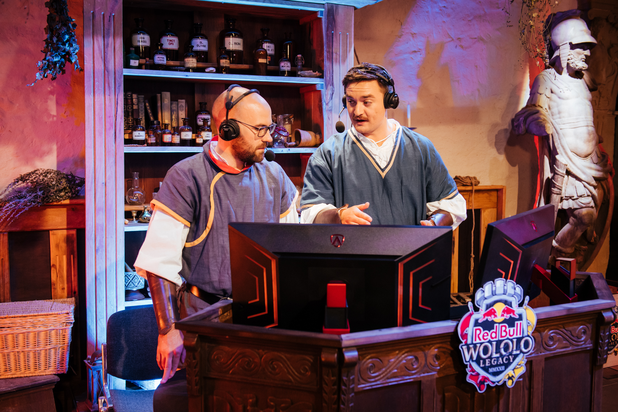 Two esports commentators in medieval-style clothing speak into headsets behind desks with gaming monitors at a Red Bull Wololo event. In the background, shelves hold bottles and a statue, setting the scene for this unique Red Bull Wololo Legacy broadcast.