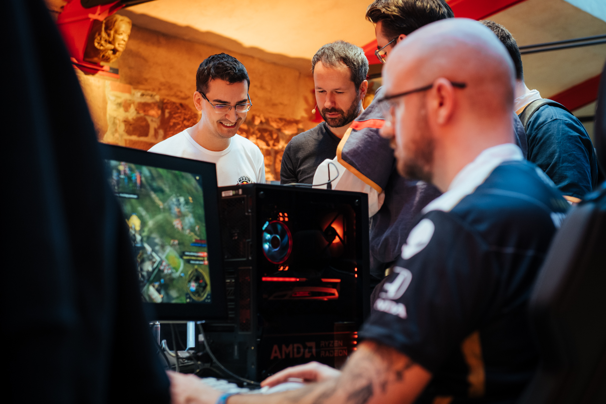 A group of people watch intently as a person plays a video game on a desktop computer at a Red Bull Wololo gaming event, with colorful lights and gaming equipment visible.