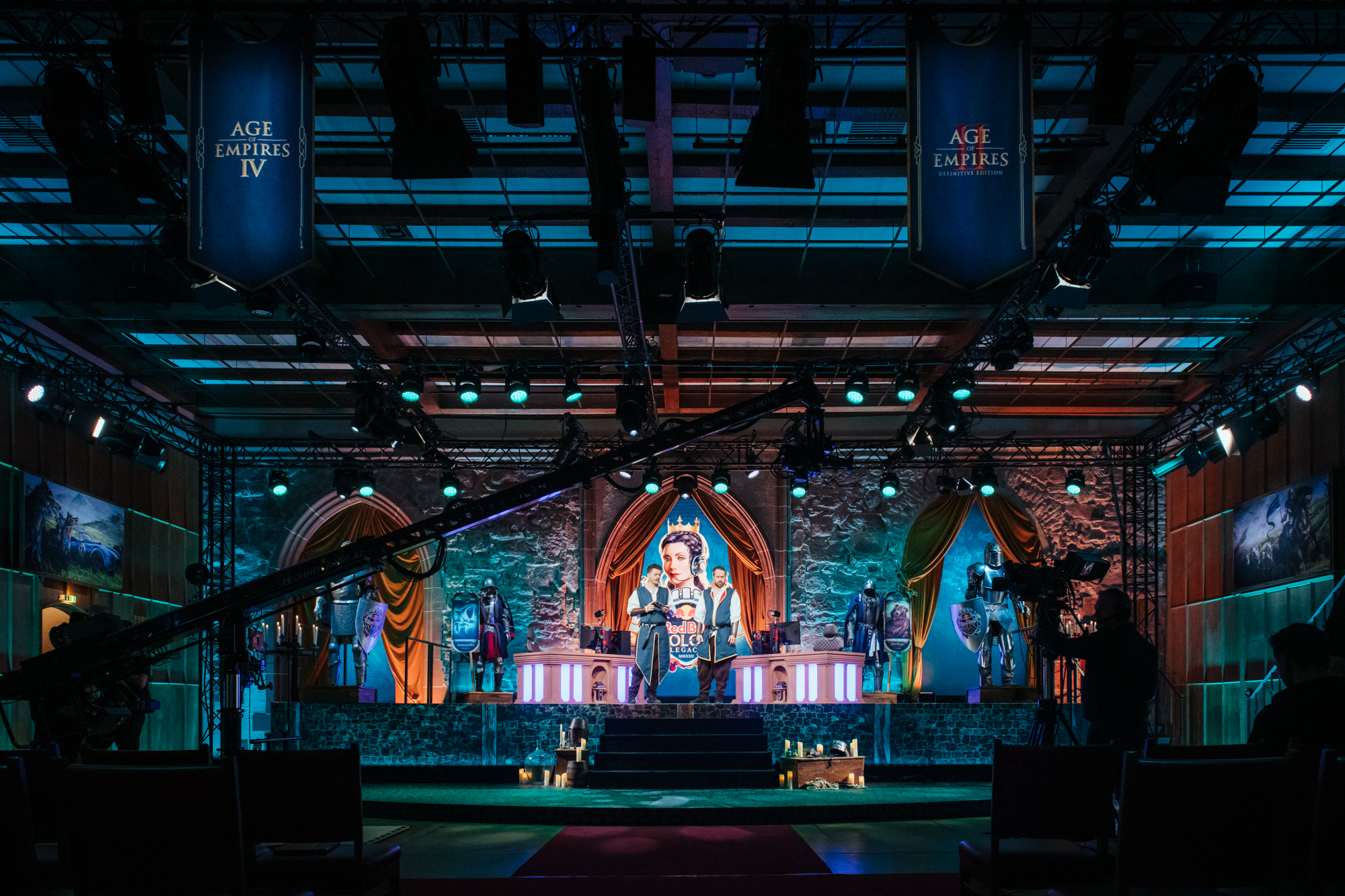 A stage set for a Red Bull Wololo Age of Empires IV event features medieval-themed decor, dramatic lighting, and a large central screen displaying a character. Cameras and crew are visible, with banners hanging overhead.