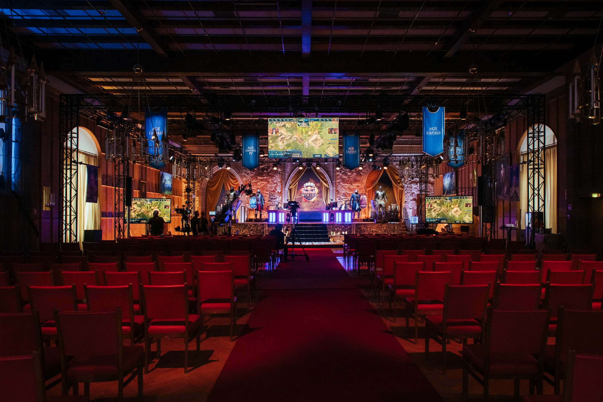 A dimly lit esports event hall with rows of red chairs faces a stage. Multiple screens showcase Red Bull Wololo, colorful lights illuminate the stage, and blue banners hang from the ceiling, creating a competitive gaming atmosphere.