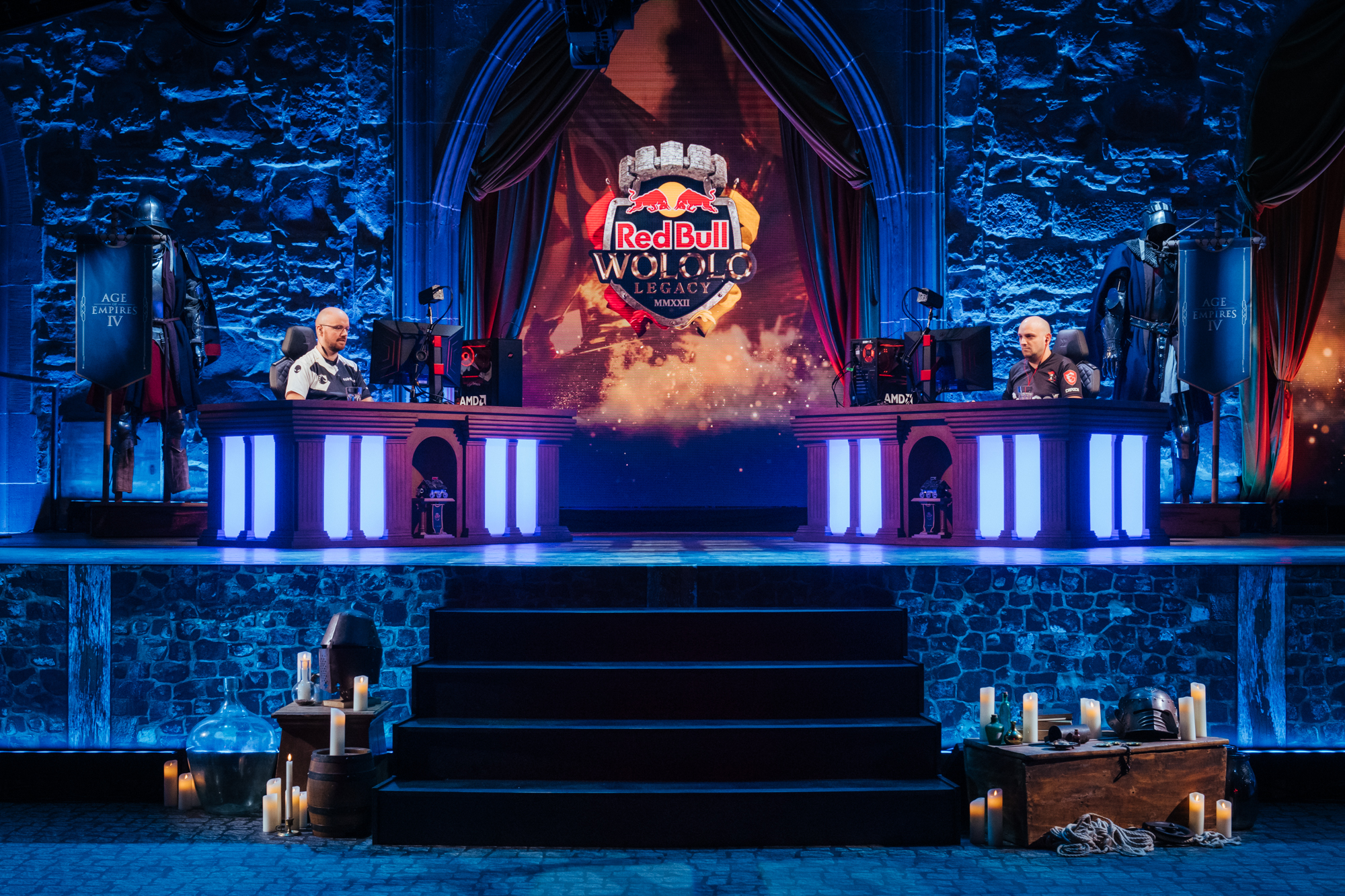 Two players compete at illuminated gaming desks on a medieval-themed stage, with Red Bull Wololo branding displayed behind them. Candles, shields, and decorative props set the scene for this epic Red Bull Wololo esports tournament atmosphere.