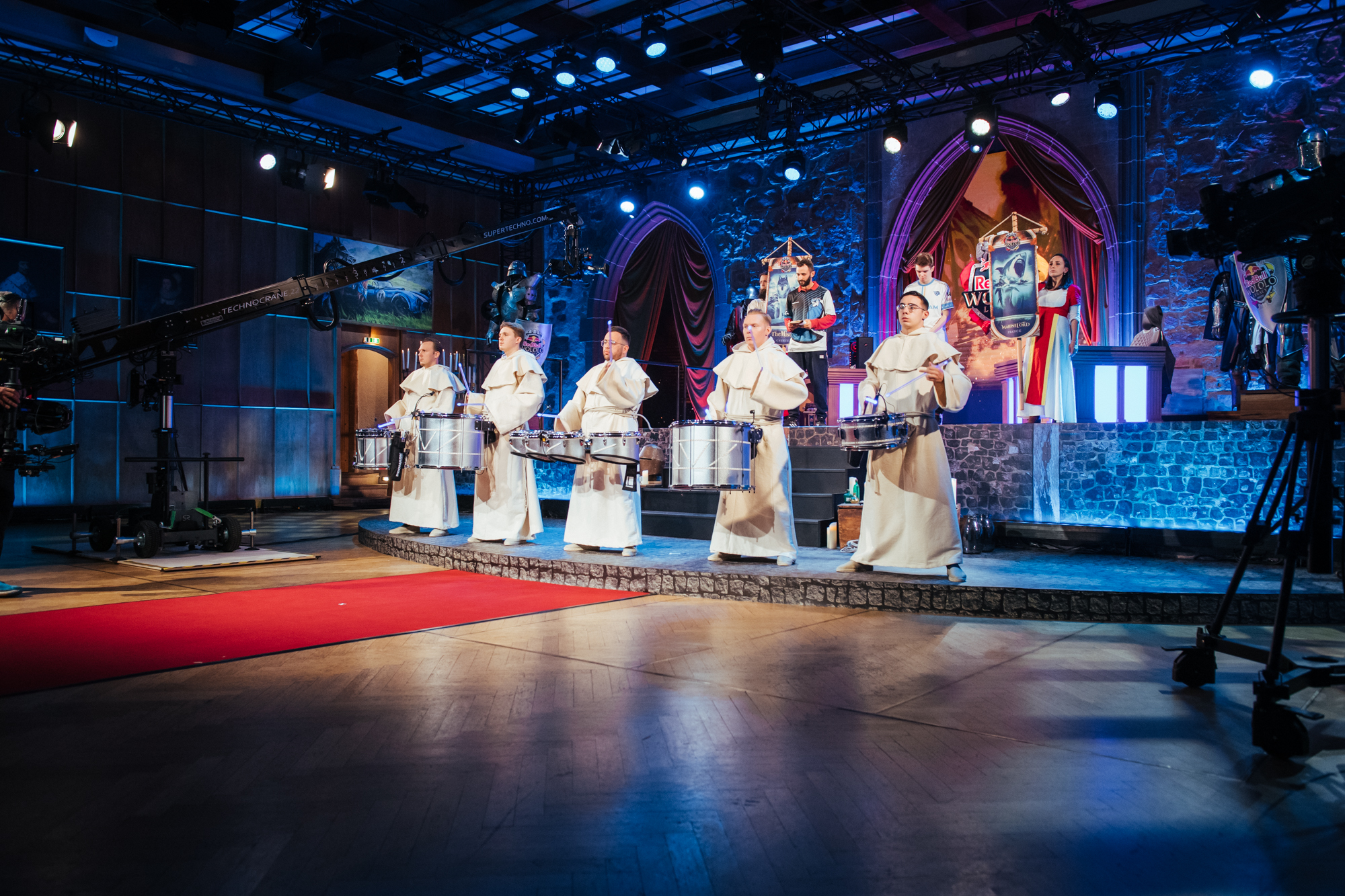 Five drummers in white robes perform on stage under blue lighting at a Red Bull Wololo event, with cameras and stage equipment visible, and a red carpet leading towards the musicians. The backdrop includes arched decorations and stone-like walls.