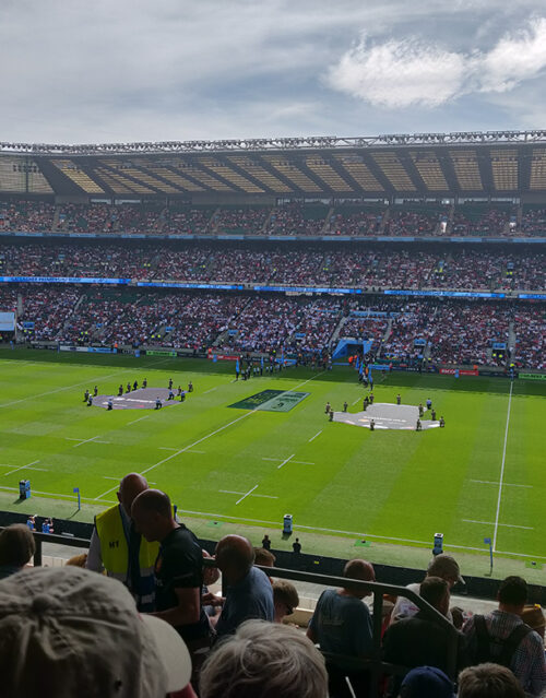 A crowded rugby stadium during daylight, with spectators in the stands and teams preparing on the field. Large banners, marketing team members, and officials are visible on the green pitch under a partly cloudy sky.
