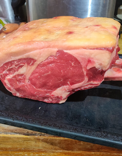 A large, raw cut of bone-in beef rib roast with a thick layer of fat on top sits on a dark cutting board, ready for the marketing team to photograph for an enticing new campaign.