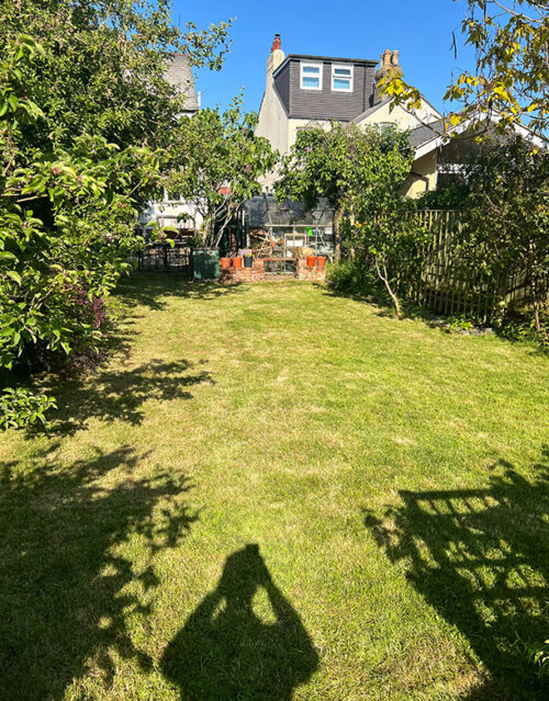 A sunlit garden with a mowed lawn, surrounded by trees and bushes, leads to a house and greenhouse in the background. The shadow of a marketing team member holding up a camera is visible in the foreground.