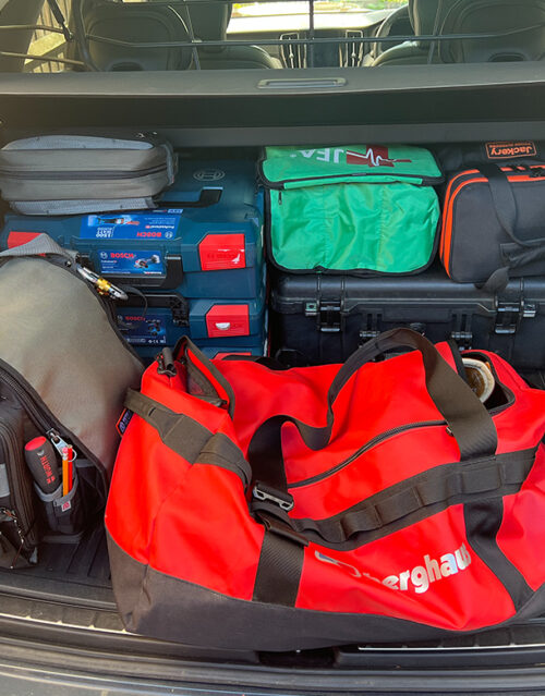 The trunk of a car filled with various bags and cases, including a large red Berghaus duffel bag—a standout in the 2024 trend review—toolboxes, a green medical kit, and other black and grey gear bags stacked neatly inside.