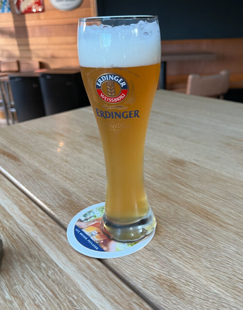 A tall glass of Erdinger beer with a foamy head sits on a coaster on a wooden table, waiting to be enjoyed by the marketing team in a casual indoor setting.