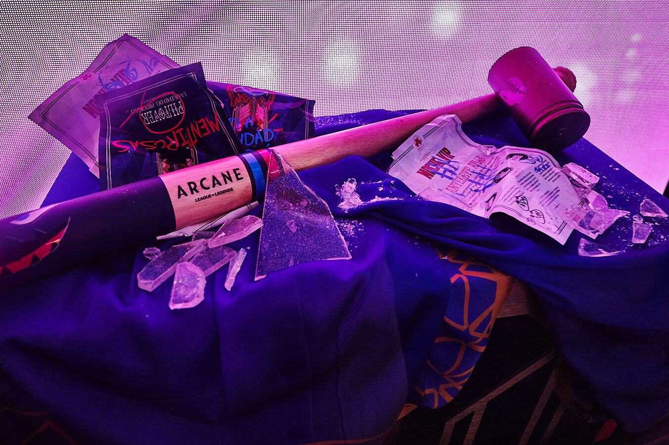 A large mallet labeled “ARCANE” lies on purple fabric amid broken glass, Netflix Arcane event cards, confetti, and a poster, all set against a glowing pink and purple background.