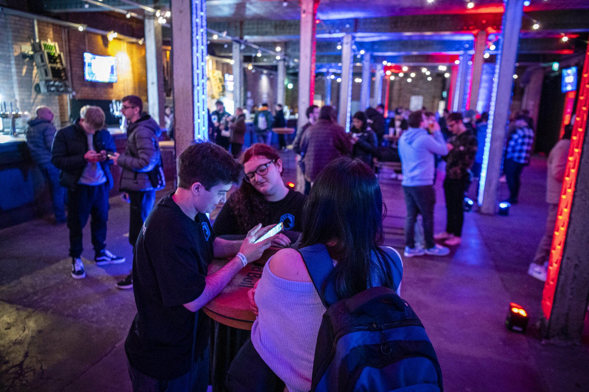 A group of people socialize in a dimly lit bar with colorful blue and red lights, some standing around high tables, while others check their phones or chat about the latest Red Bull Home Ground action in the background.