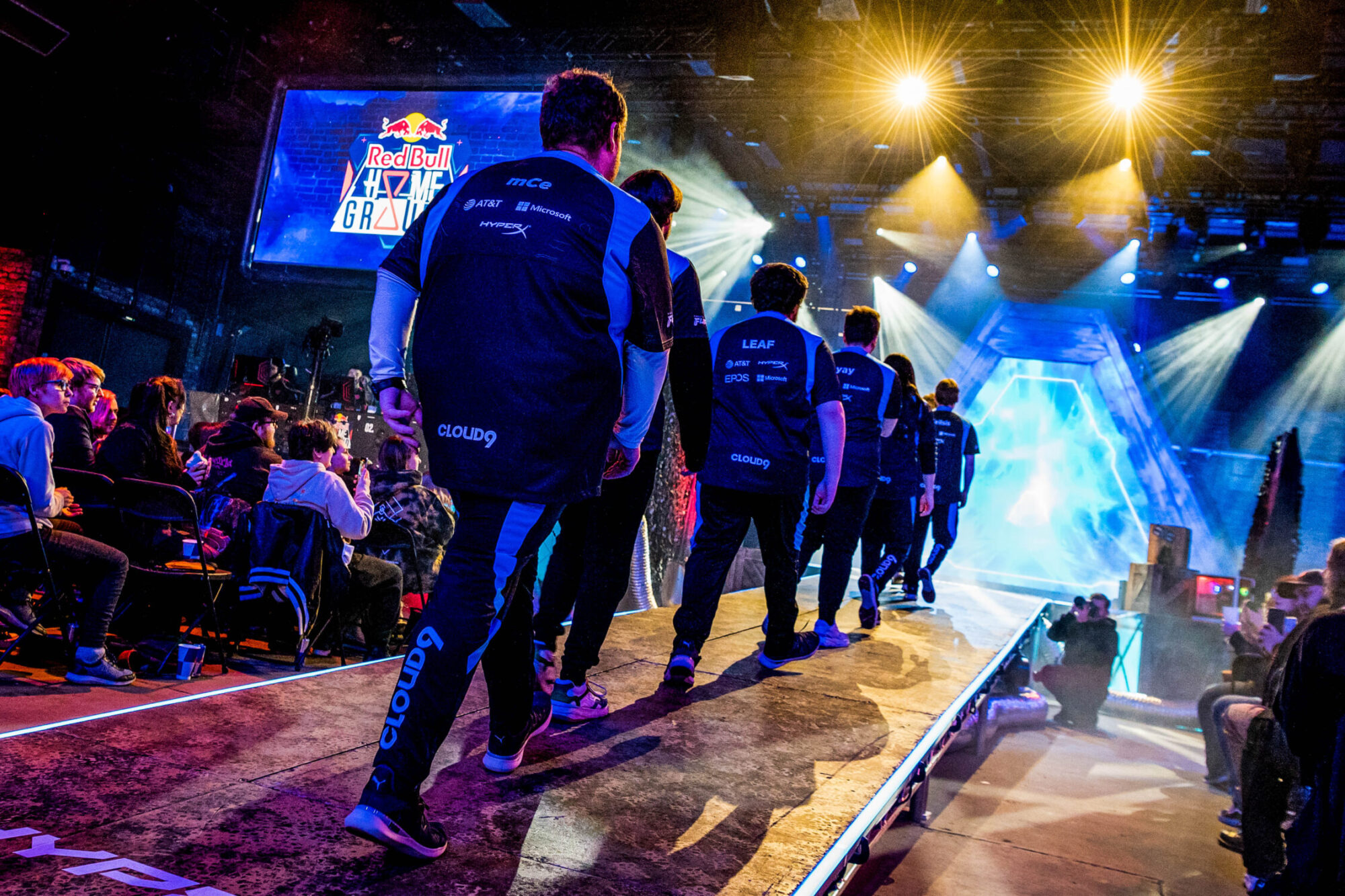 A team of esports players wearing matching jerseys walks onstage at a gaming event, with an audience on both sides and colorful stage lights illuminating the venue during the Red Bull Home Ground tournament.