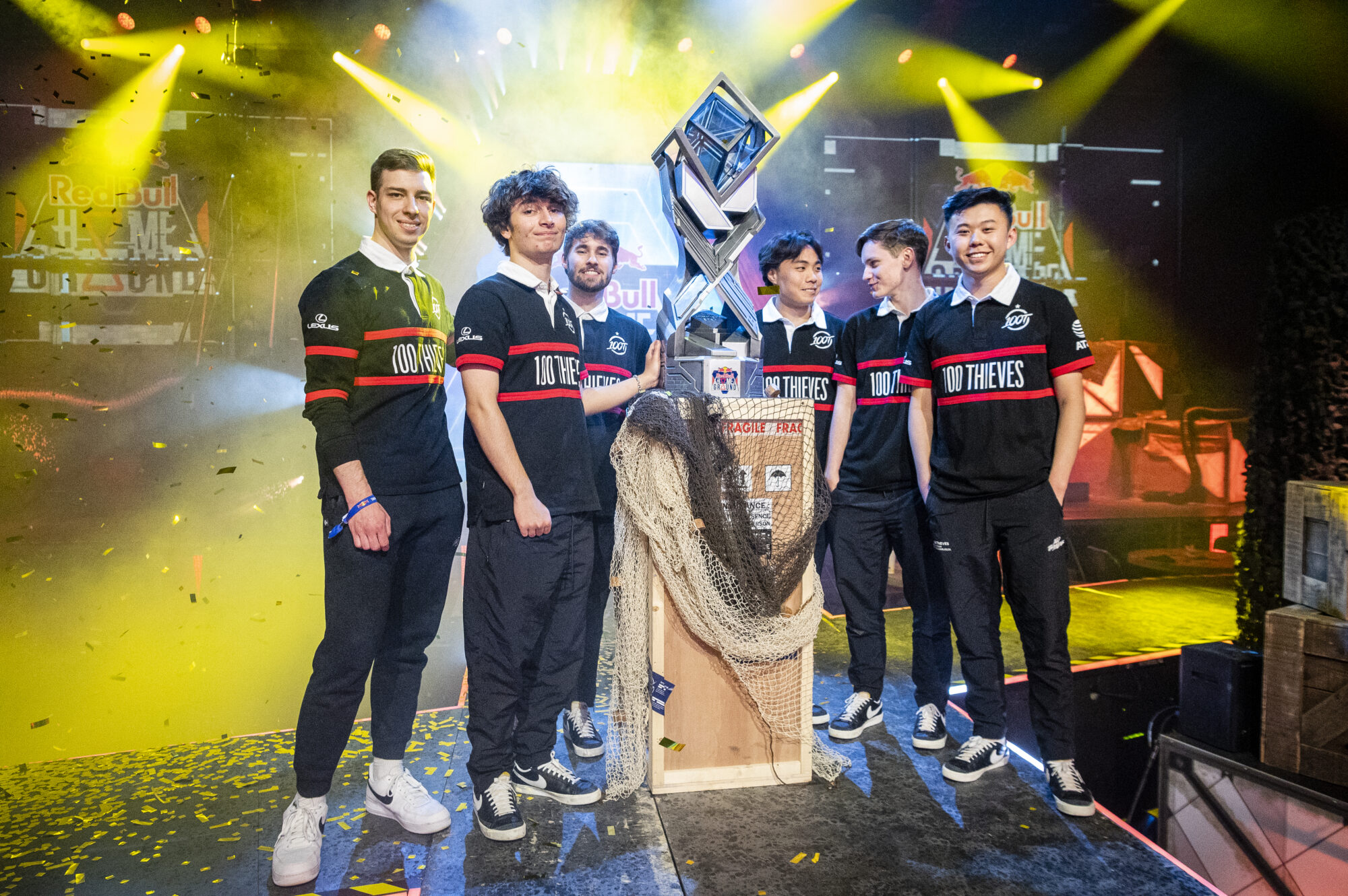 Six young men in matching black 100 Thieves jerseys stand smiling around a large trophy on stage. Colorful lights, confetti, and the Red Bull Gaming logo celebrate their Red Bull Home Ground victory in the background.