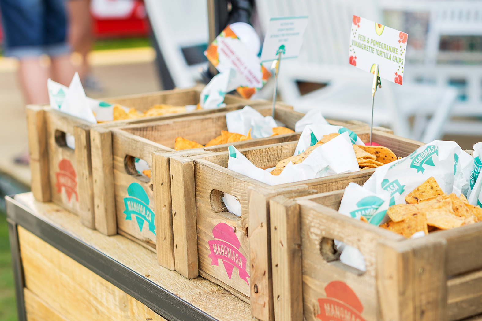 Wooden crates filled with tortilla chips lined up on a table, each labeled with colorful stickers—reminiscent of a Manomasa case study. Small signs describe each crate, and white napkins are tucked in with the chips.