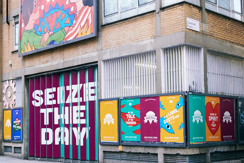 Storefront with large text SEIZE THE DAY on striped panels, colorful posters with motivational phrases, and a mural above showing cartoon faces and hands—reminiscent of the vibrant style found in the Manomasa case study. Brick walls and barred windows complete the look.