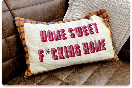 A rectangular pillow with a checkered border sits on a brown leather couch. The contact collider of bold style and humor is clear in its red text: “HOME SWEET F*CKING HOME,” with the “U” replaced by an asterisk.