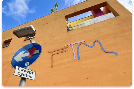 A blue one-way traffic sign with a white arrow and a sticker stands below an Except cycles sign, creating a visual contact collider against the orange wall’s abstract line art and the modern building rising above.