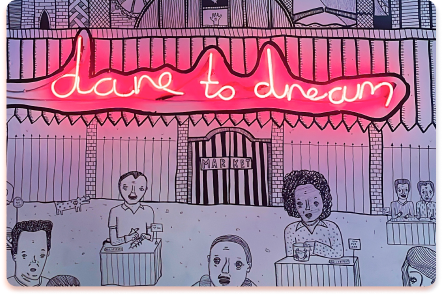 A neon sign reading dare to dream glows above a black-and-white drawing of people in contact at tables outside a market, with stylized buildings in the background, creating a lively urban collider of ideas.