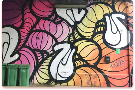 Colorful mural with abstract, swirling shapes in pink, yellow, and orange tones on a brick wall. Three green recycling bins are lined up in front of the artwork, creating a contact collider of color and function. A small graffiti tag sits on the right.