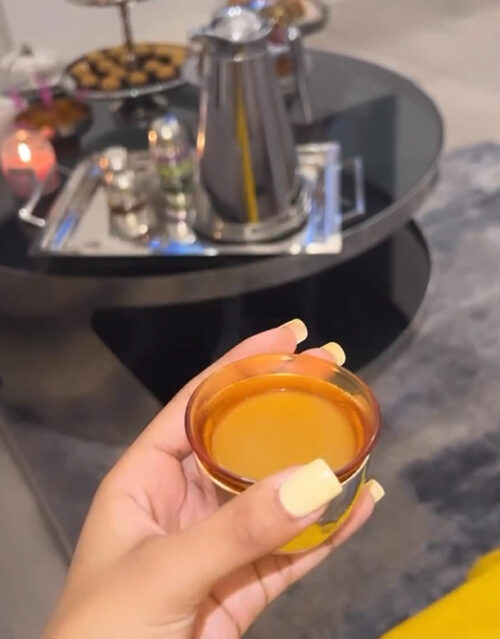 A person with manicured nails holds a small glass cup of tea. In the background, a marketing team gathers around a round black coffee table with a metal teapot, food trays, and snacks arranged on it.
