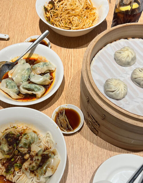 A wooden table set with plates of dumplings in chili sauce, a bowl of noodles, a steamer basket with soup dumplings, a small dish of soy sauce, and a glass of soda—perfect for sharing with your marketing team.