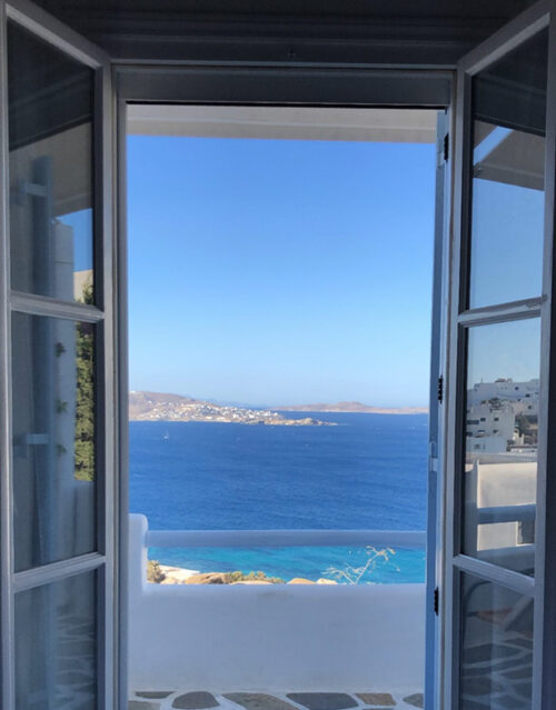 Open double doors reveal a balcony where a marketing team can unwind, taking in the stunning view of the deep blue sea, white buildings, and distant hills under a clear sky.