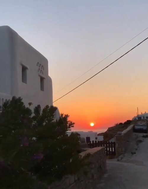 A white building with simple, boxy architecture stands near a narrow street as the sun sets over the sea, casting an orange and pink glow. Along the roadside, some cars and plants are visible—perhaps even where a marketing team gathers after work.