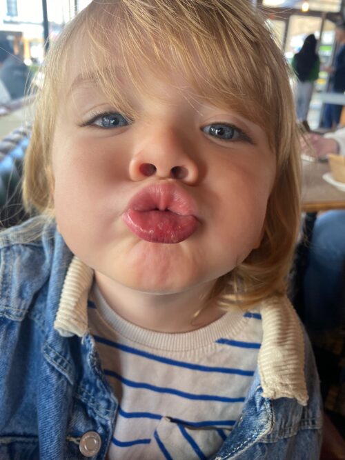 A young child with blond hair puckers their lips for a kiss, wearing a striped shirt and denim jacket, indoors at what appears to be a café or restaurant—perhaps capturing the charm your marketing team is aiming for.