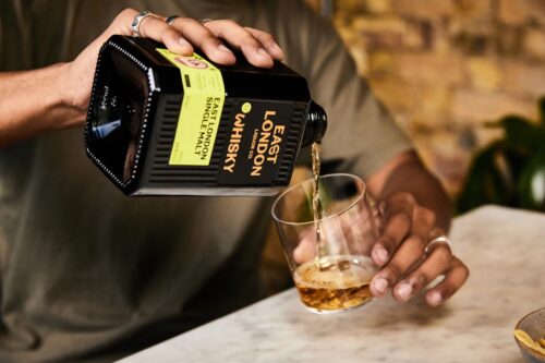 A member of the marketing team pours East London Whisky from a black bottle into a glass tumbler on a marble table, with a blurred brick wall and greenery in the background.