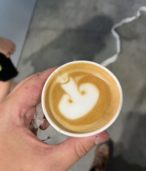 A hand holds a cup of coffee with latte art resembling an abstract shape, possibly a mushroom or heart, on a light brown foam background—perfect for energizing the marketing team. The scene is set against a gray floor.