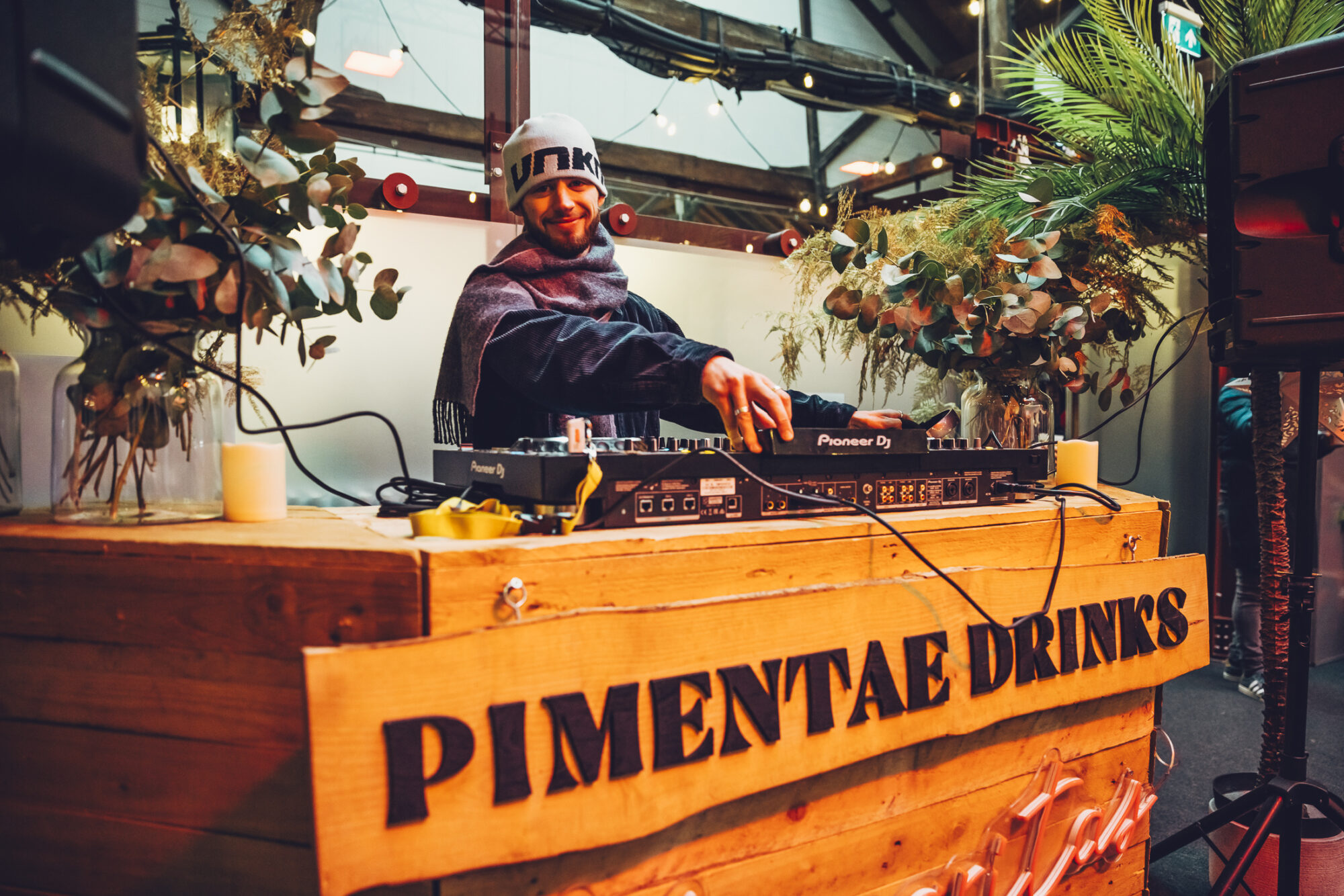 A DJ wearing a gray beanie and scarf stands behind a wooden booth labeled PIMENTAE DRINKS, mixing music on DJ equipment at the Waitrose Winter Festival. The booth is decorated with greenery and set in an indoor venue with string lights.