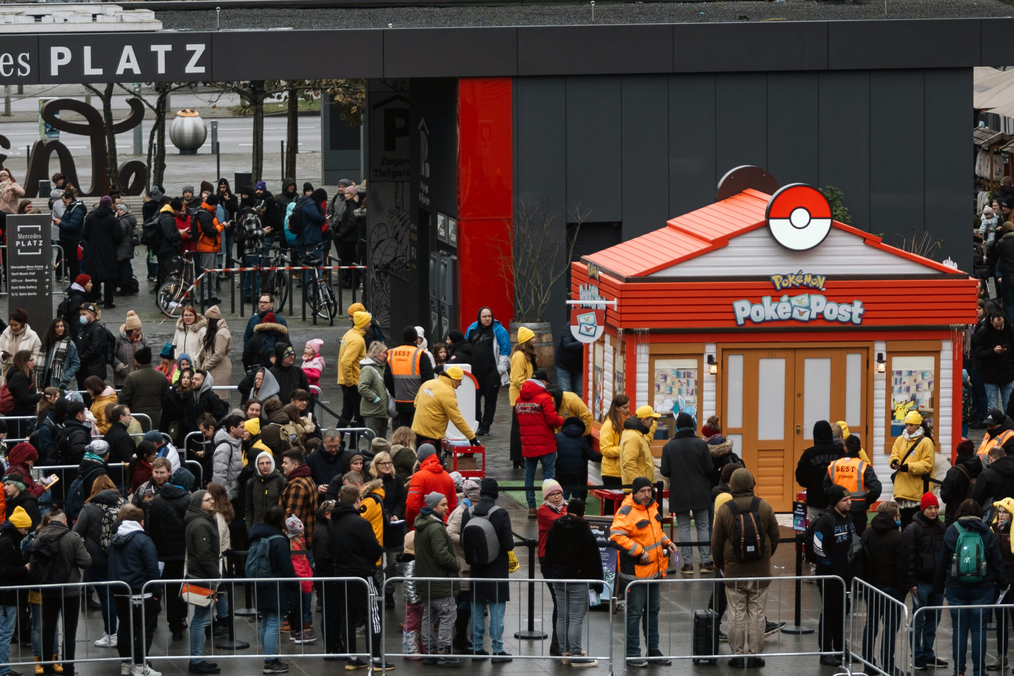 A large crowd waits in line outside a Pokémon Poke Post kiosk, with staff in yellow jackets assisting. The scene is set in a city plaza on a cloudy day, with barriers neatly guiding eager fans toward the themed kiosk.