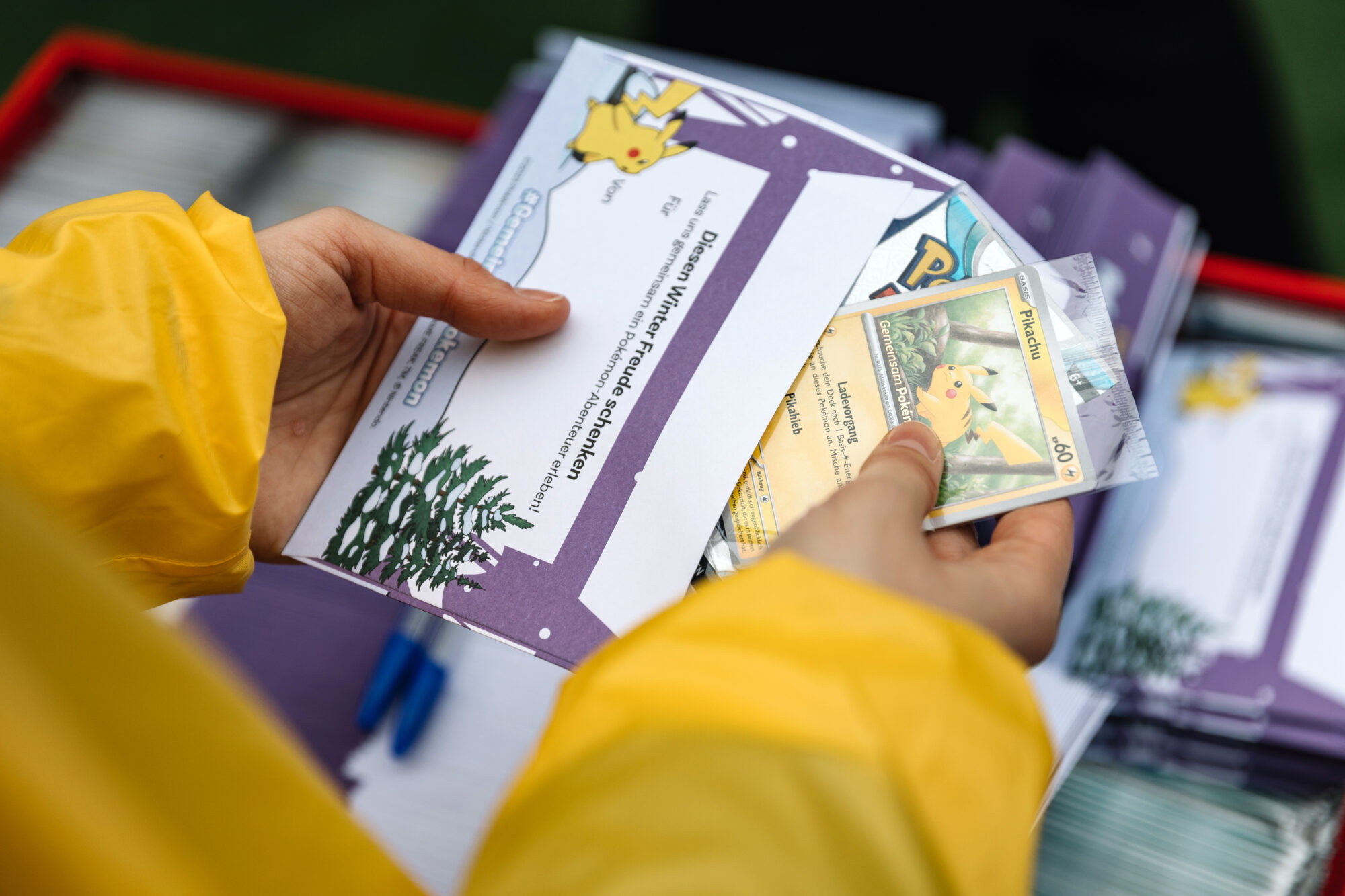 A person in a yellow jacket holds a Pokémon Poke Post folder with a Pikachu card and holiday-themed materials, including a pine tree card and printed text. Similar folders are stacked nearby.