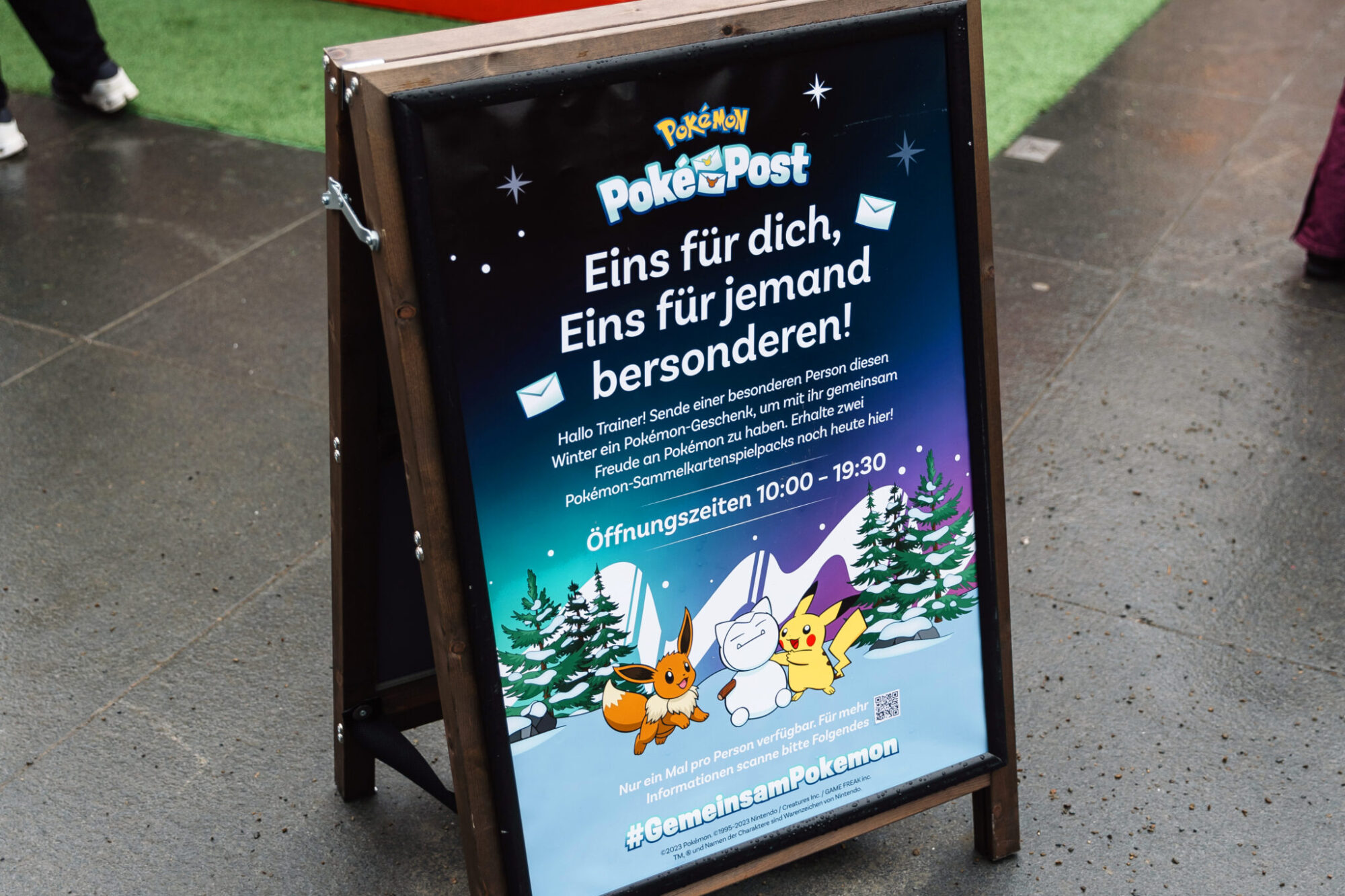A wooden sidewalk sign displays a winter-themed Pokémon Poke Post poster with German text, featuring illustrations of Pikachu, Alolan Vulpix, and Eevee with snow and gifts, on a wet pavement near green artificial grass.