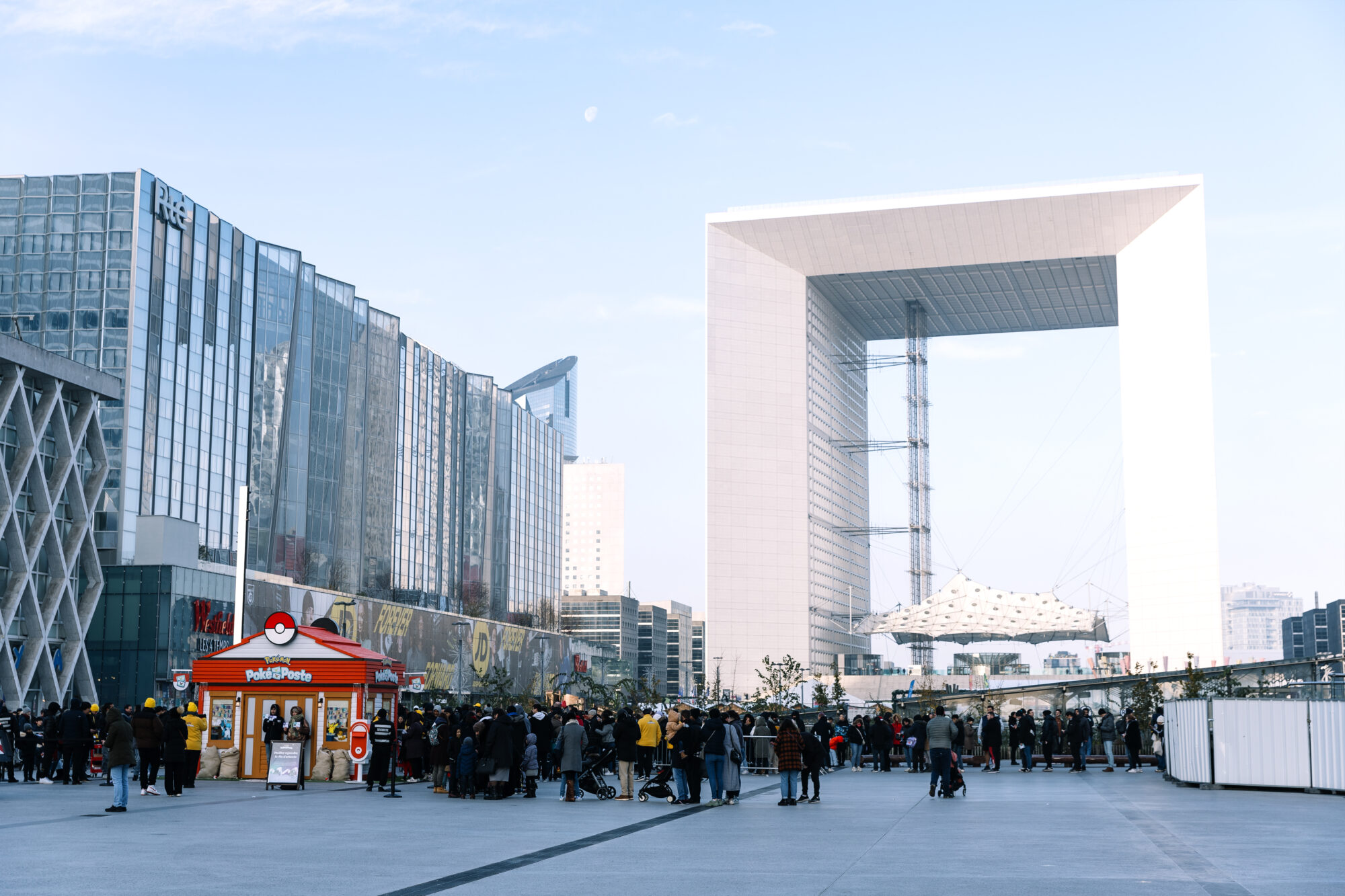Large modern plaza with a crowd near a red and white Pokémon Poke Post kiosk; tall glass buildings and the distinctive square arch of La Défense, Paris, stand in the background under a clear sky.