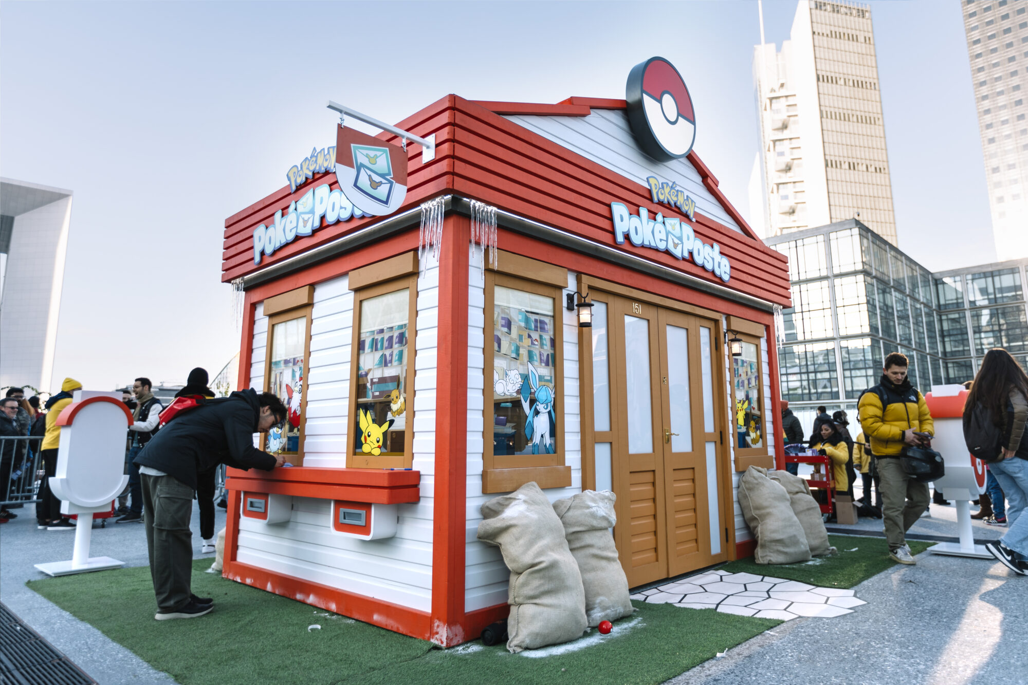 A small Pokémon Poke Post kiosk shaped like a house, featuring Poké Ball decorations and Pokémon images, stands outdoors. People gather around it, with tall modern buildings rising in the background.