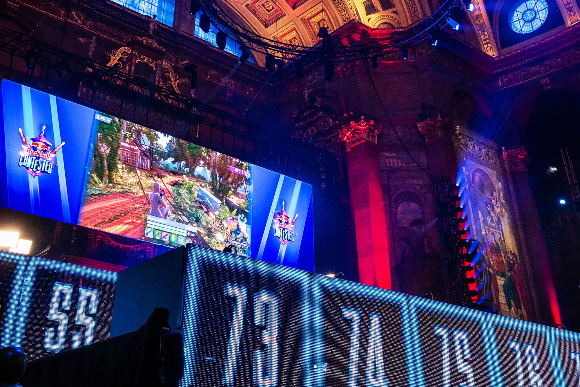 A large gaming event stage with ornate architecture, illuminated by blue and red lights. A big screen displays a video game from Red Bull Contested, while numbered booths for players are visible below the screen.