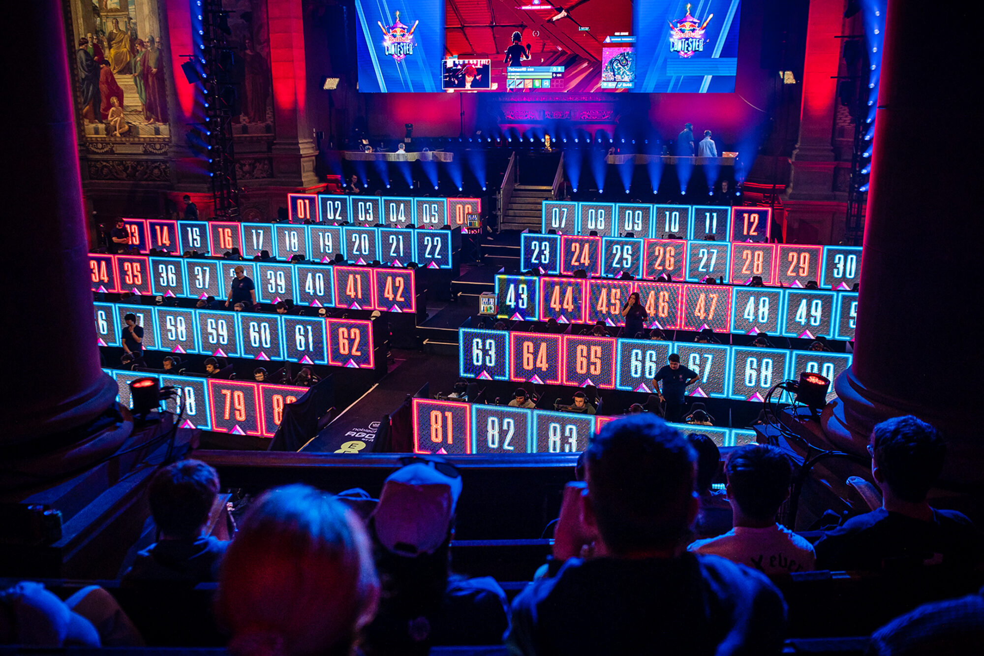 A large esports event, Red Bull Contested, features multiple digital screens displaying numbers across the stage as players compete on an elevated platform. An audience watches from the foreground in a dimly lit, vibrant arena.