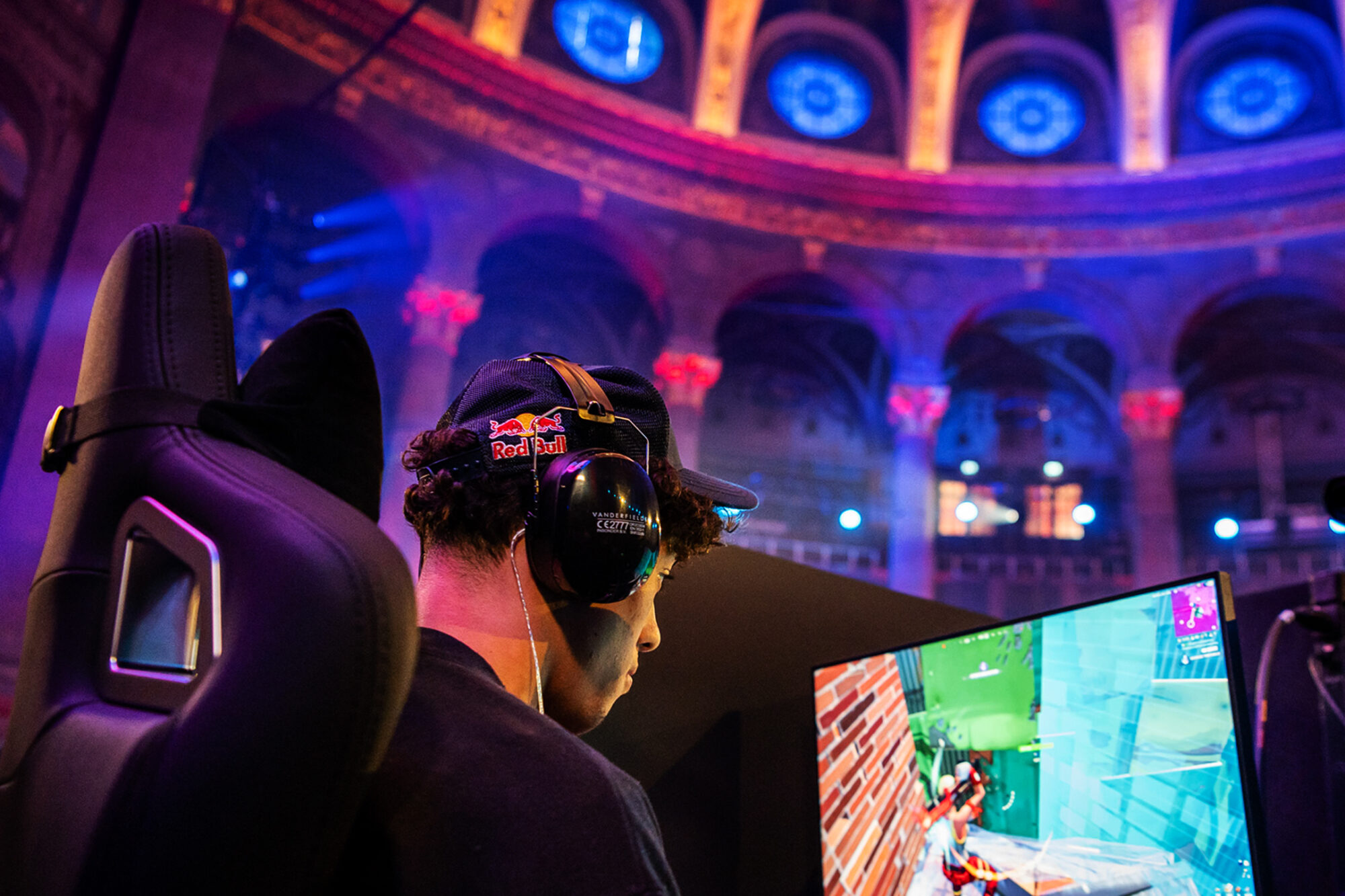 A gamer wearing headphones and a Red Bull cap sits in front of a monitor, immersed in a video game at the vibrant Red Bull Contested arena, featuring colorful lighting and striking architectural arches in the background.
