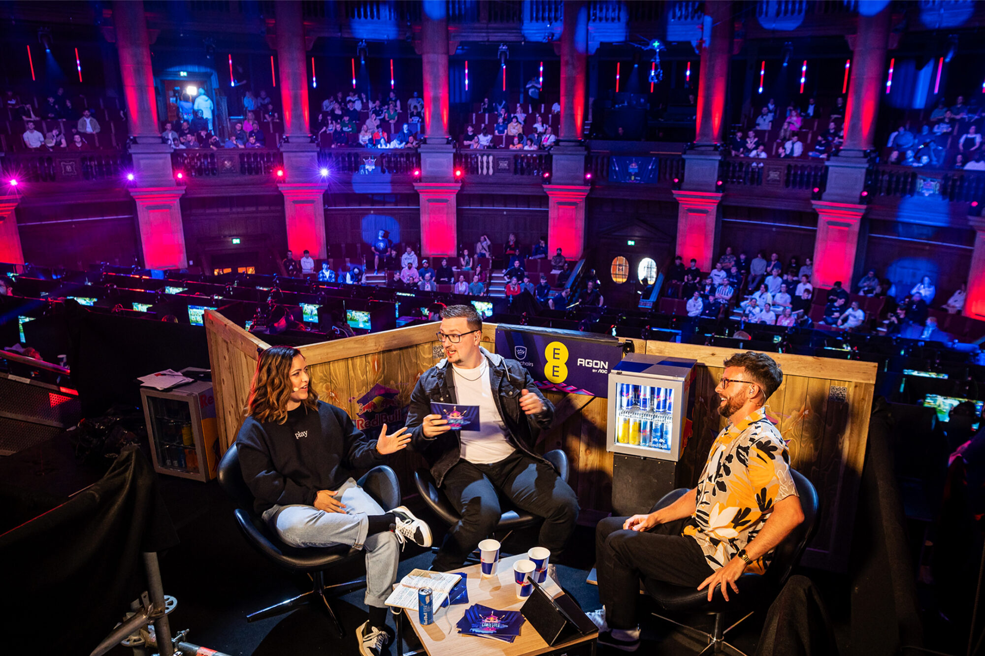 Three people sit and chat on a stage set with drinks and microphones at Red Bull Contested, facing an audience in a large, colorful hall lit with blue and red lights. Rows of computers and spectators fill the background.