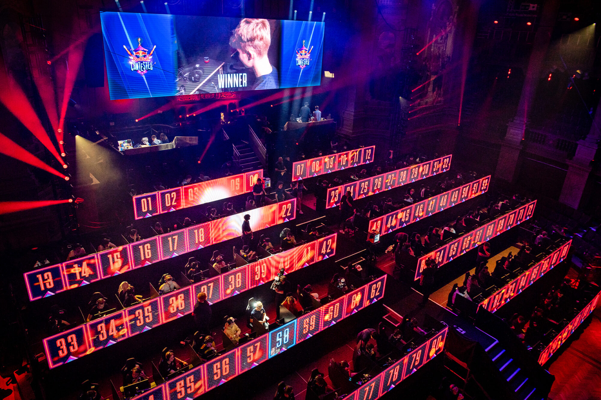 A large esports event, Red Bull Contested, features rows of players at glowing numbered computer stations, red and blue stage lighting, and a “WINNER” screen above with the champion’s photo as spectators watch from the surrounding area.