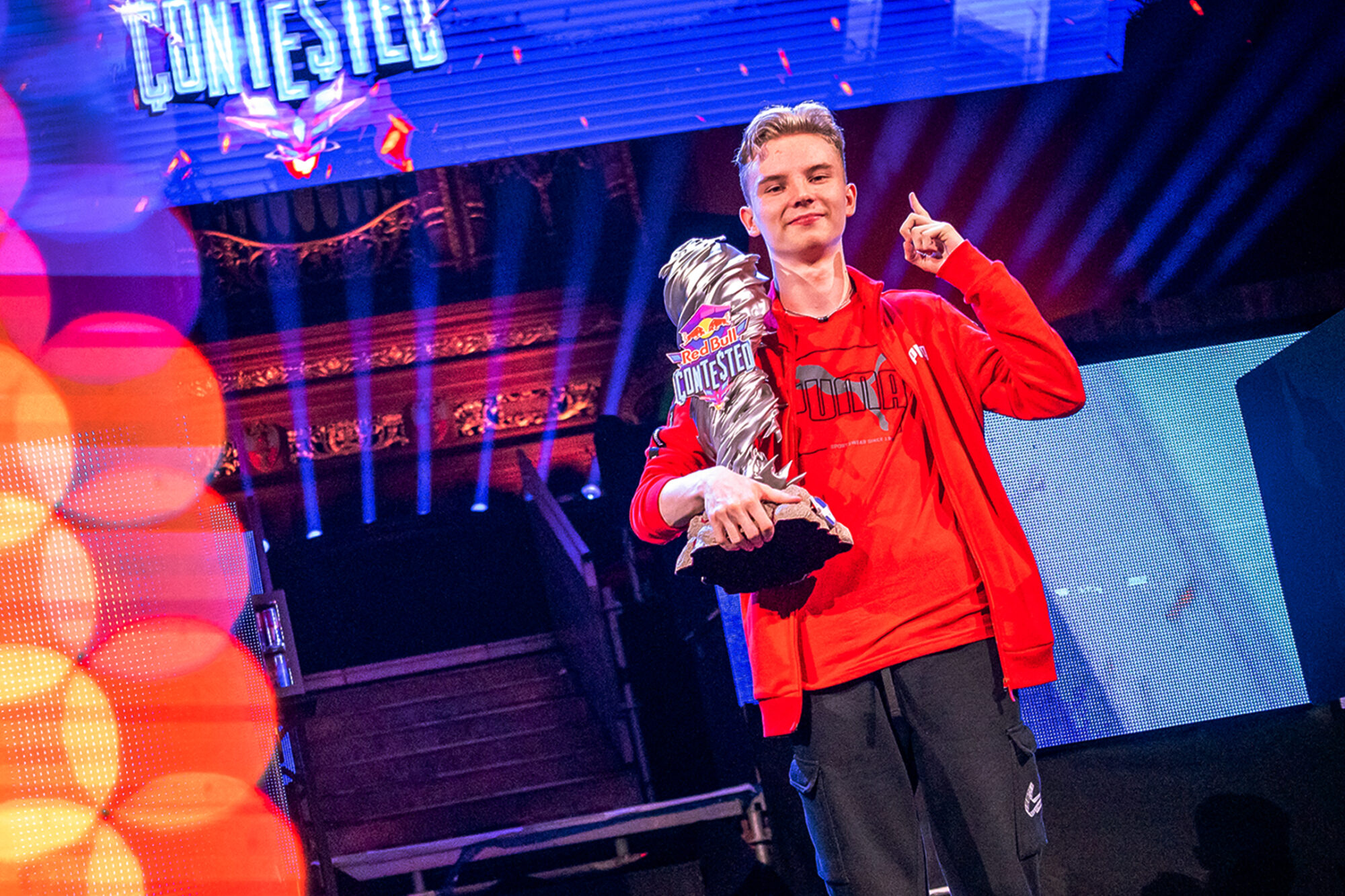 A young man in a red jacket holds a large trophy and points upward, standing on stage with colorful lights and a glowing Red Bull Contested sign above him. The atmosphere is energetic and celebratory.