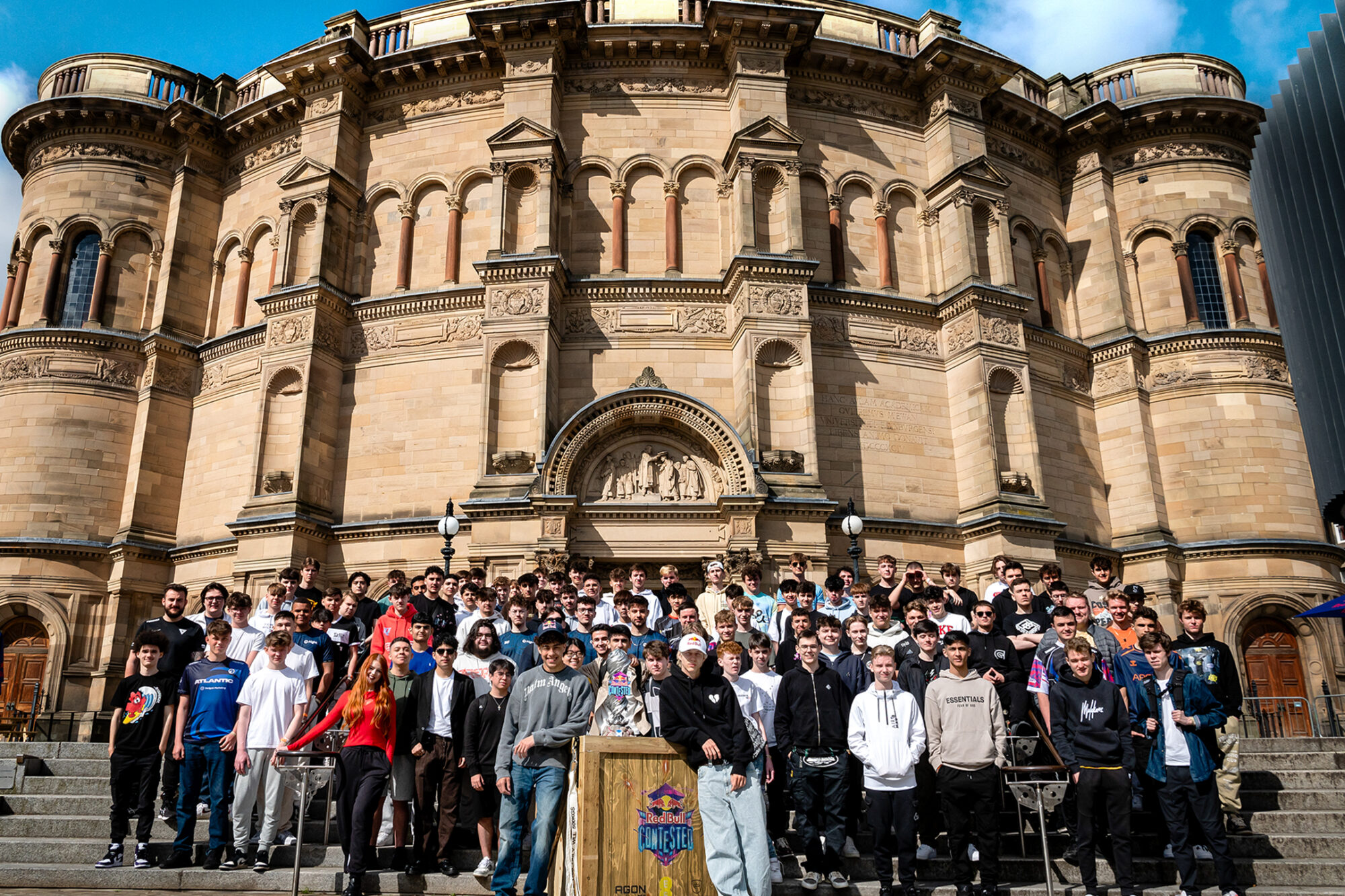 A large group of students stands on the steps of a grand, historic stone building with arched windows and decorative carvings, posing for a group photo under a partly cloudy sky after attending the Red Bull Contested event.
