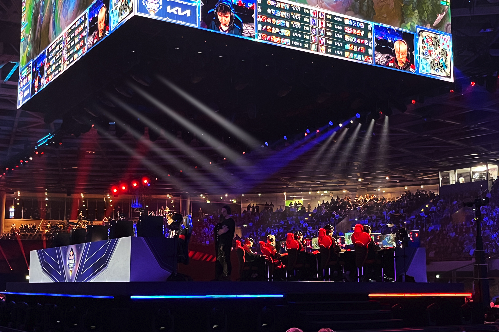 A large esports arena buzzes with excitement as teams compete on stage at computer desks. A massive screen displays player stats and the Red Bull LOIO logo, while a crowd of spectators watches from the stands.