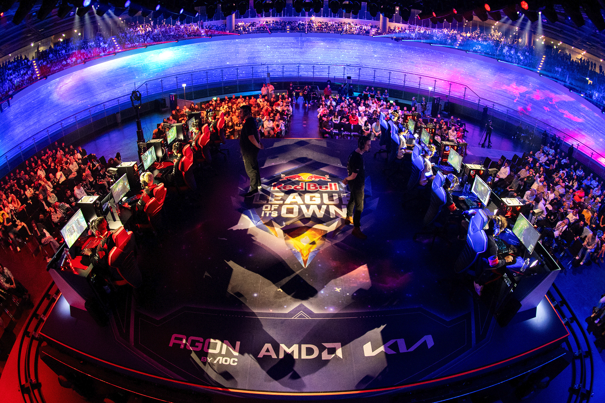 A large esports arena filled with spectators surrounds two teams of players at computer stations. The floor displays the Red Bull LOIO logo, with colorful lighting and sponsor banners illuminating the vibrant venue.