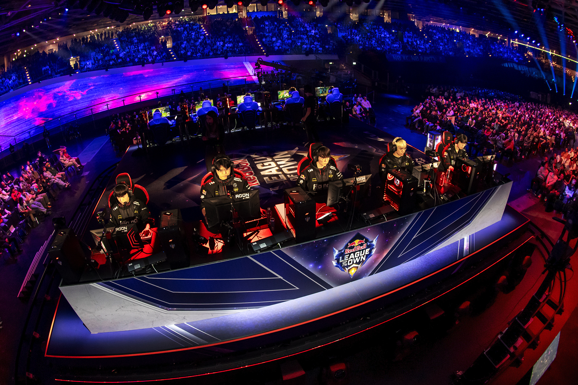 A professional esports team plays at a row of computers on a brightly lit stage during the Red Bull LOIO League of Legends tournament, with a large live audience and colorful lighting in the background.
