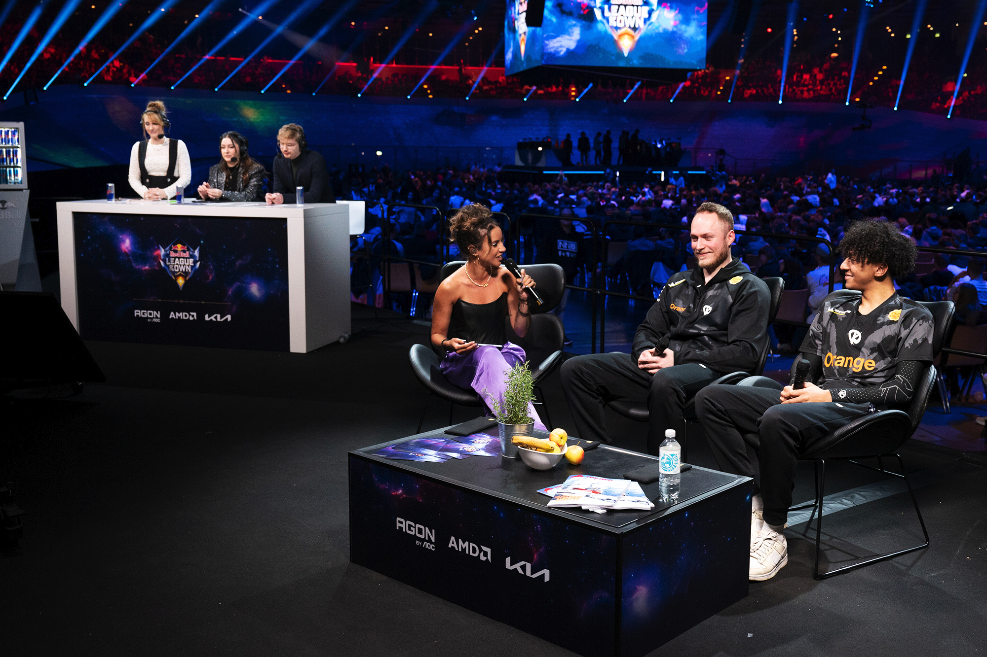 A live esports event with two players on a couch being interviewed by a host, while panelists sit at a desk in the background. The stage is lit with colorful lights, and Red Bull LOIO sponsor logos are visible.