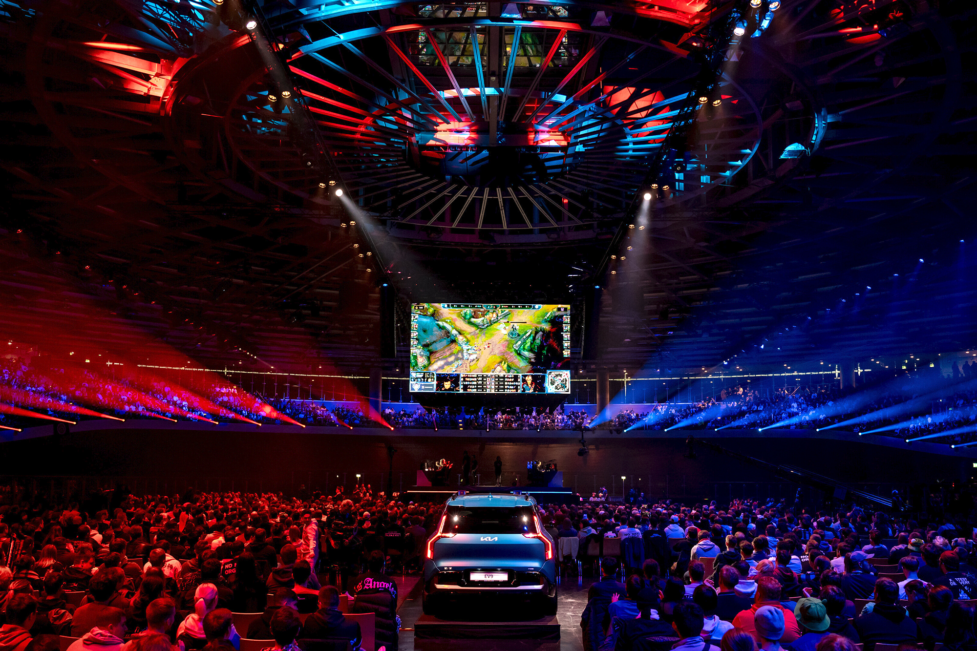 A large audience watches an esports event in a vibrant, colorful arena with blue and red lighting, a car on stage, and a giant screen displaying the Red Bull LOIO video game competition.
