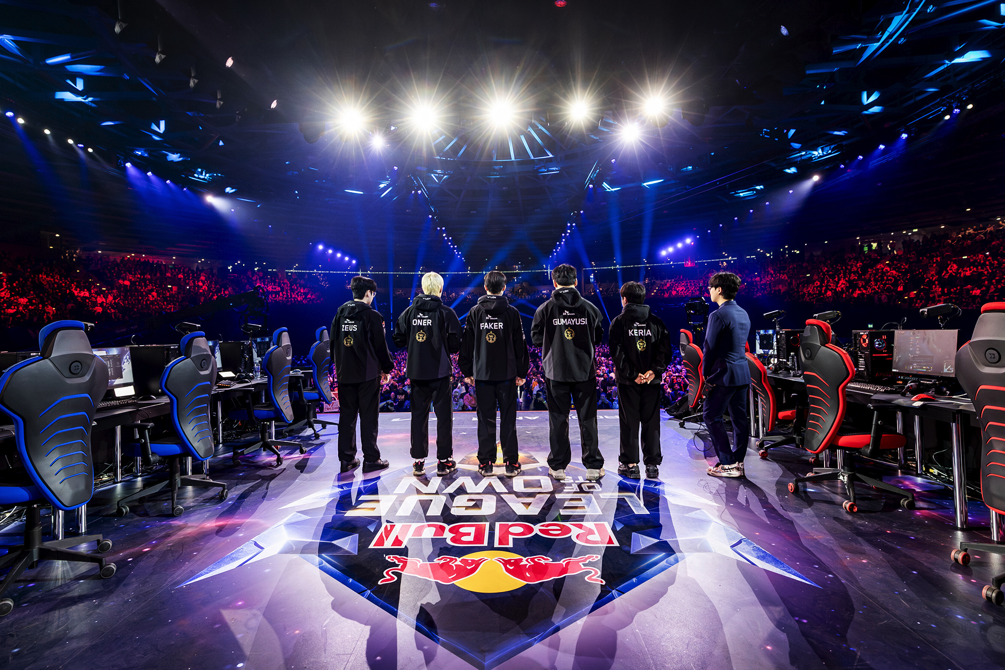 Six esports players stand on a brightly lit stage facing a large, cheering audience, with Red Bull LOIO branding on the floor and gaming chairs and computers on each side.