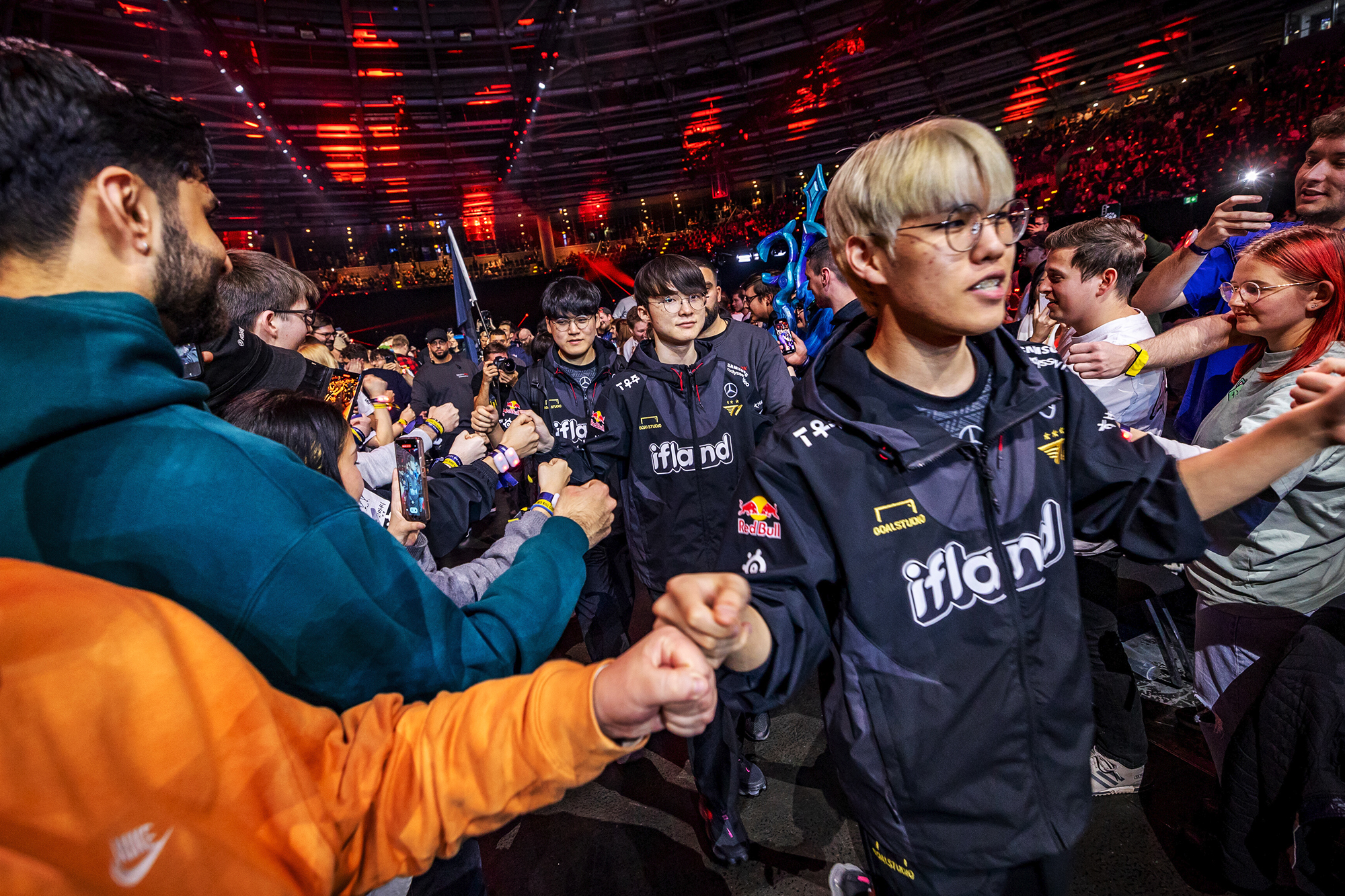 A group of esports players in matching Red Bull LOIO jerseys walk through a cheering crowd in an arena, exchanging fist bumps with fans under colorful lights.